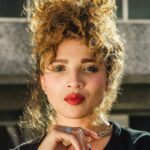 Portrait of a young woman with curly hair and red lips wearing a necklace outdoors.